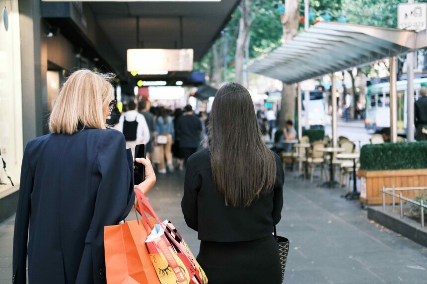 Two shoppers walk along a busy city shopping street carrying retail bags, with cafés, pedestrians and public transport visible in the background.