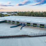Aerial view of a modern distribution centre with multiple loading docks, a large concrete hardstand and surrounding industrial precinct, bordered by trees under a partly cloudy sky.