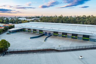 Aerial view of a modern distribution centre with multiple loading docks, a large concrete hardstand and surrounding industrial precinct, bordered by trees under a partly cloudy sky.