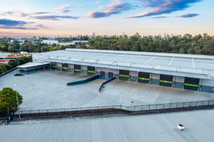 Aerial view of a modern distribution centre with multiple loading docks, a large concrete hardstand and surrounding industrial precinct, bordered by trees under a partly cloudy sky.