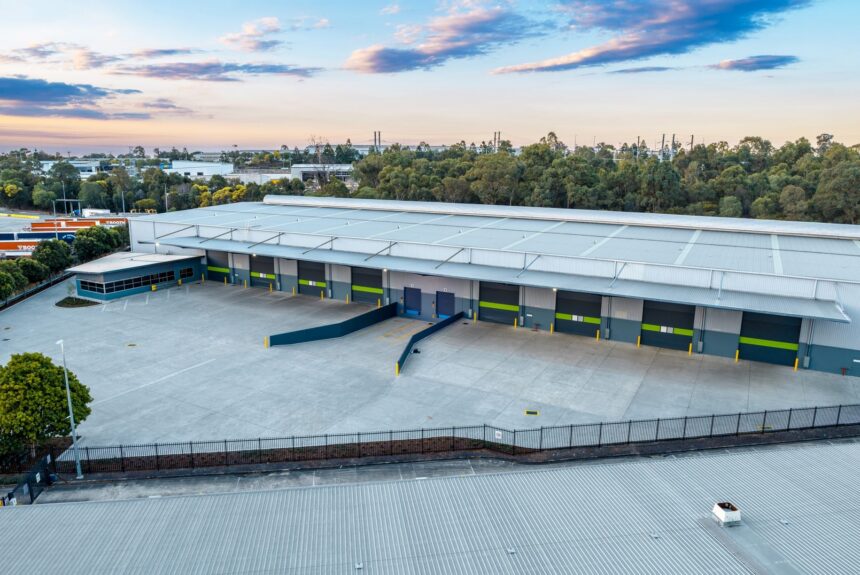 Aerial view of a modern distribution centre with multiple loading docks, a large concrete hardstand and surrounding industrial precinct, bordered by trees under a partly cloudy sky.