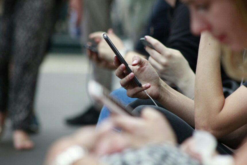 Close-up of people sitting side by side using smartphones, with multiple hands holding mobile devices, illustrating social media and digital connectivity.