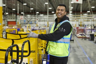 A smiling Amazon warehouse worker wearing a high-visibility vest beside fulfilment equipment.
