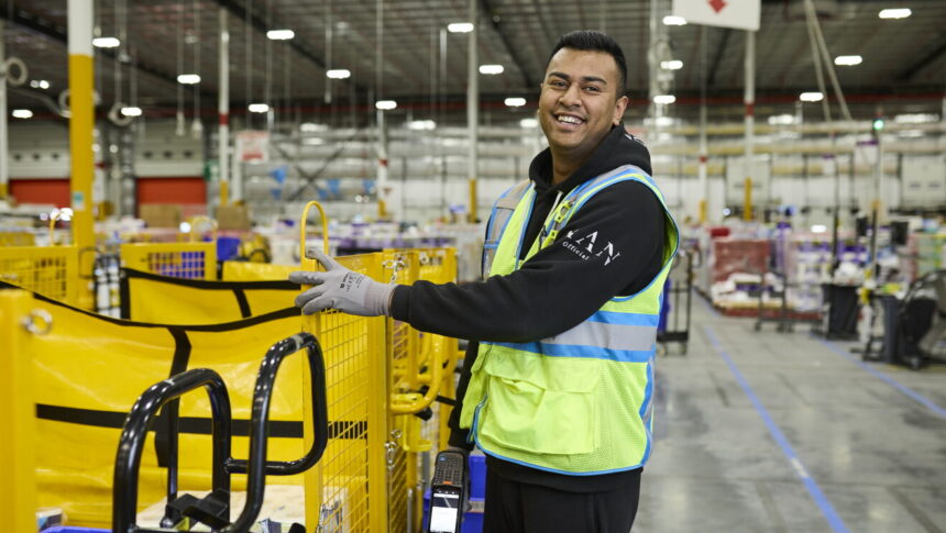 A smiling Amazon warehouse worker wearing a high-visibility vest beside fulfilment equipment.