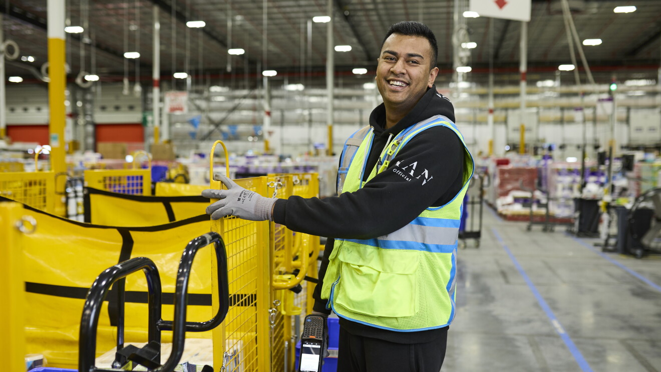 A smiling Amazon warehouse worker wearing a high-visibility vest beside fulfilment equipment.