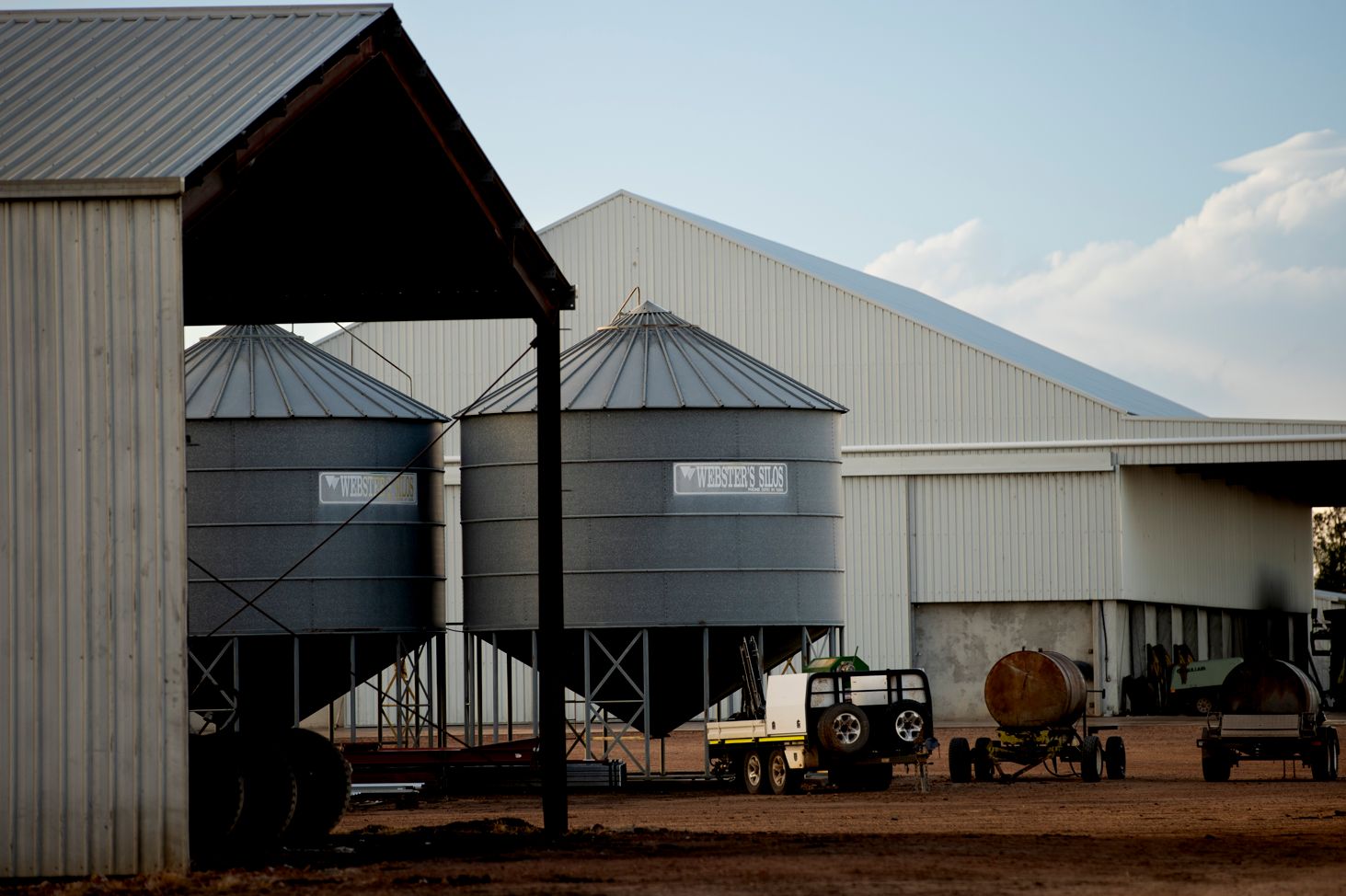 Grain storage silos and large sheds at an agricultural production facility, with farm equipment and trailers nearby.