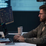 Man working at a computer workstation with multiple screens displaying cybersecurity monitoring and data dashboards.