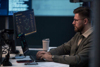 Man working at a computer workstation with multiple screens displaying cybersecurity monitoring and data dashboards.