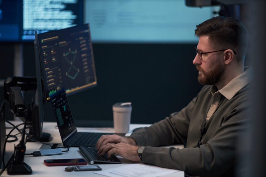 Man working at a computer workstation with multiple screens displaying cybersecurity monitoring and data dashboards.