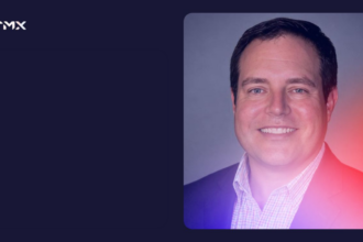 Professional headshot of a smiling man wearing a dark blazer and checked shirt, featured in a TMX-branded graphic with a navy background and pink and purple gradient.