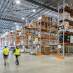 Two workers in high-visibility vests walking through a large warehouse fitted with Colby selective pallet racking, stocked with pallets and cardboard boxes.