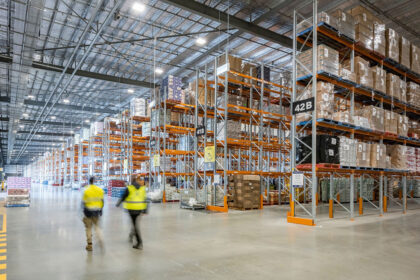 Two workers in high-visibility vests walking through a large warehouse fitted with Colby selective pallet racking, stocked with pallets and cardboard boxes.