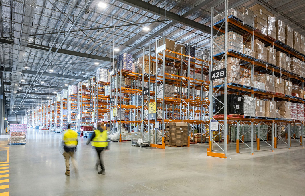 Two workers in high-visibility vests walking through a large warehouse fitted with Colby selective pallet racking, stocked with pallets and cardboard boxes.