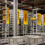 Automated warehouse storage and conveyor systems at a DHL Health Logistics facility, featuring high-density shelving, grey storage containers and DHL-branded signage.