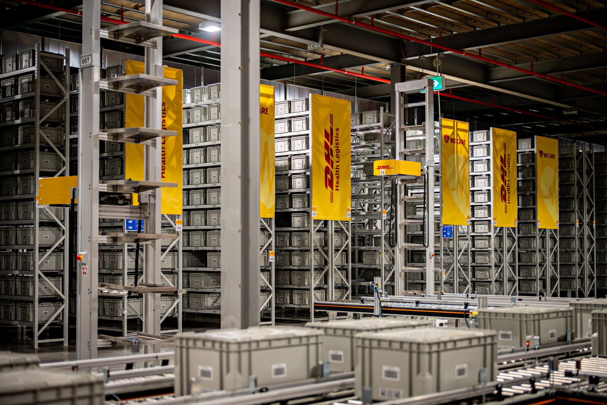 Automated warehouse storage and conveyor systems at a DHL Health Logistics facility, featuring high-density shelving, grey storage containers and DHL-branded signage.