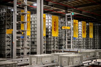 Automated warehouse storage and conveyor systems at a DHL Health Logistics facility, featuring high-density shelving, grey storage containers and DHL-branded signage.