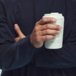 Person wearing a navy jumper holding a light grey made by Fressko reusable coffee cup, with the brand's logo visible on the front.