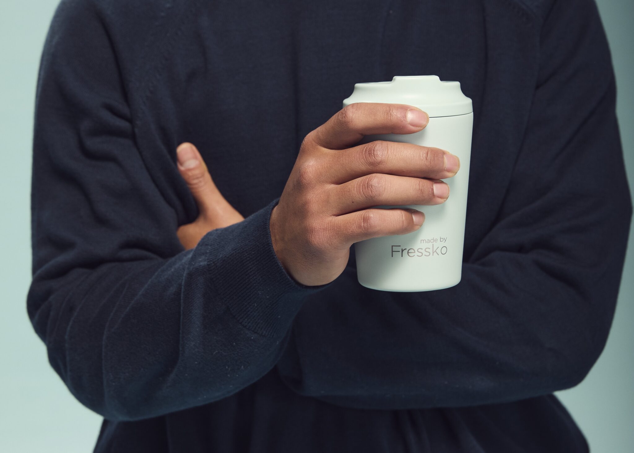 Person wearing a navy jumper holding a light grey made by Fressko reusable coffee cup, with the brand's logo visible on the front.
