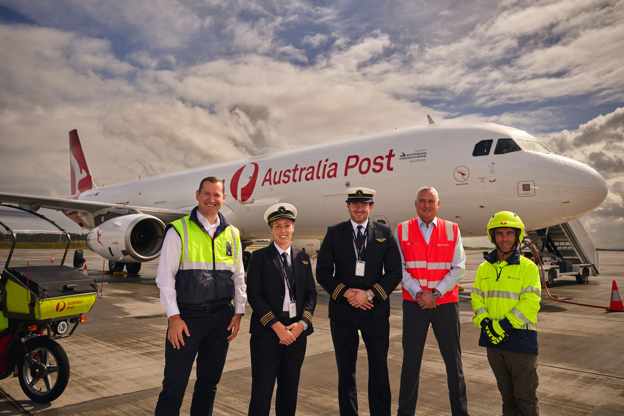 Qantas Freight and Australia Post representatives standing alongside pilots and ground crew in front of an Australia Post-branded Airbus A321 freighter aircraft.